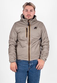 Giacca a piumino beige con cappuccio, chiusura frontale con cerniera e cerniera arancione decorativa. Presenta un logo sul petto e polsini elastici. Indossata sopra jeans blu.