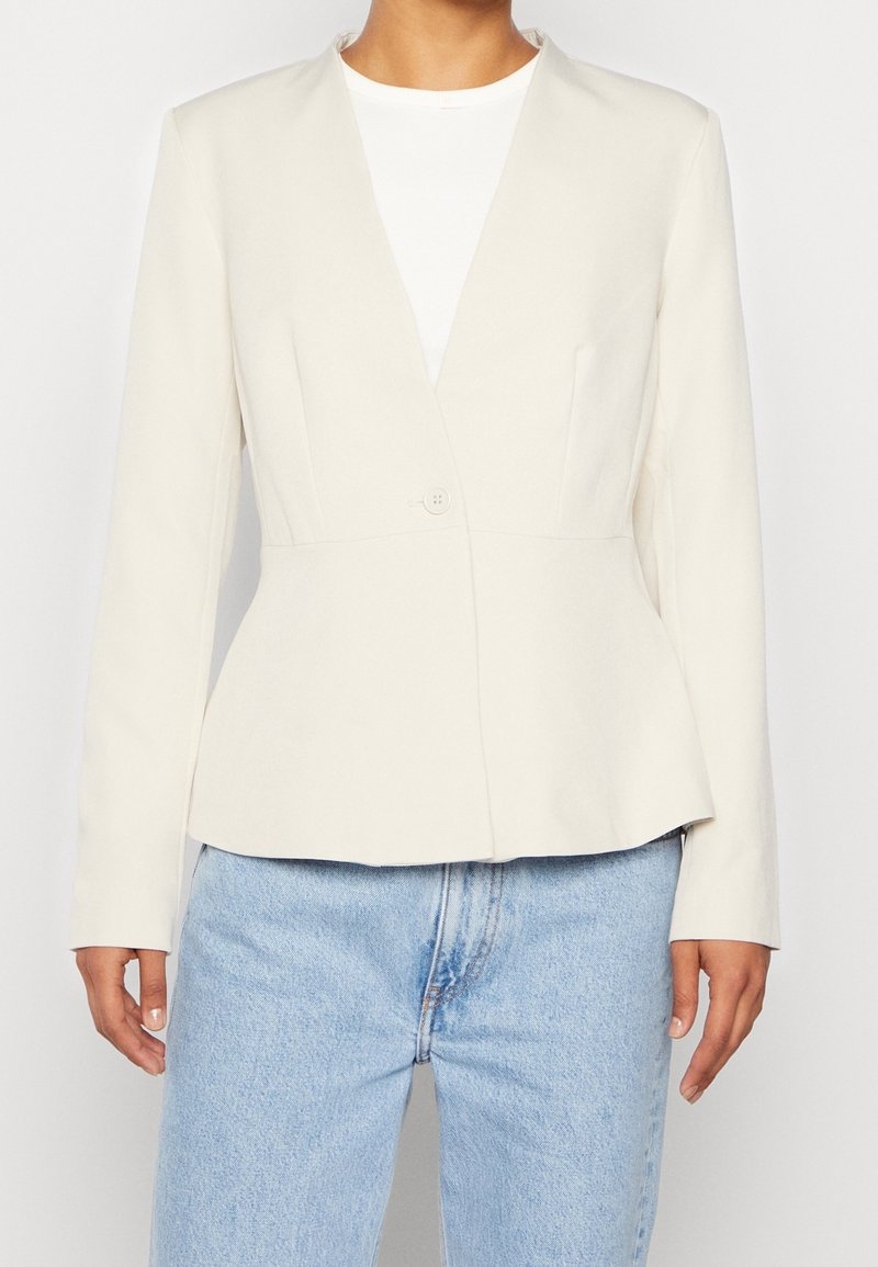 Blazer beige cintré avec un col en V, une fermeture à un bouton, une taille structurée, des manches longues et une texture de tissu lisse, associé à un jean bleu.
