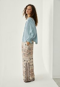 Mujer con cabello rizado que lleva una blusa bordada de color azul claro y pantalones anchos con estampado, de pie con las manos en los bolsillos, girada de lado.