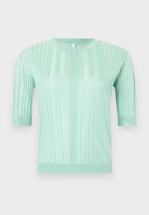 Femme portant un haut vert menthe à manches courtes, un short long argenté, des chaussures blanches, tenant un sac à main beige, debout devant un fond blanc uni.