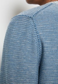 Blau strukturierter Strickartikel mit feinen Streifen und einer sichtbaren Nahtdetail an der Schulter, der eine neutrale Farbpalette präsentiert.