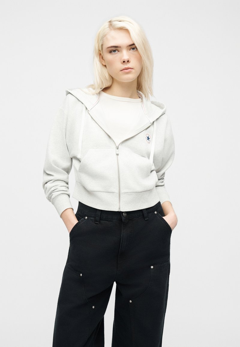 Jonge persoon met blond haar, gekleed in een lichtgrijze hoodie met rits en zwarte broek, staand met handen in de zakken tegen een effen achtergrond.