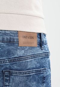 Jeans in denim blu sbiadito con un'etichetta in pelle con la scritta "ONLY & SONS" cucita sul retro. Presenta tasche posteriori e dettagli di cucitura.