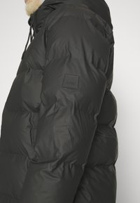 Zwarte pufferjack met een gestructureerde oppervlakte, een capuchon en een gestikt ontwerp. Opvallend rubber Rains-logo op de mouw.