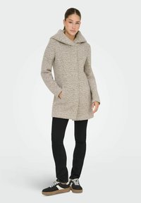 Manteau beige texturé avec une large capuche, fermeture à boutons sur le devant et poches latérales, associé à un jean noir et des baskets noires avec des accents blancs.