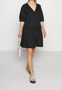 Černé bavlněné šaty s výstřihem do V a balónovými rukávy, nařaseným pasem a rozšířenou sukní, doplněné béžovými sandály na křížový pásek a psaníčkem.