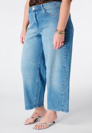 Personne portant un jean large bleu clair, un haut noir, des sandales argentées à talons et des bracelets au poignet gauche.