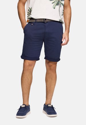 Hombre con pantalones cortos azul marino remangados, cinturón tejido, camisa de estampado tropical metida por dentro y zapatillas casuales azul marino.