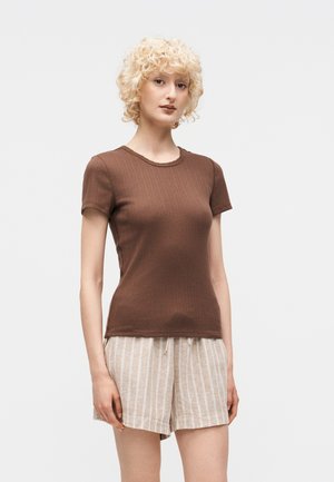 Γυναίκα με ξανθά σγουρά μαλλιά, που φοράει στενό καφέ κοντομάνικο τοπ και μπεζ ριγέ σορτς με κορδόνια, στέκεται μπροστά σε απλό φόντο.