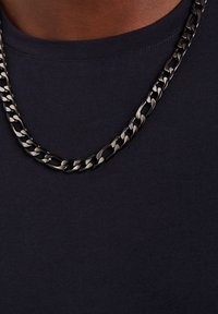 Zilverkleurige metalen ketting met grote, in elkaar grijpende schakels die op een donkerblauwe shirt ligt. Soepele textuur met een glanzende afwerking.