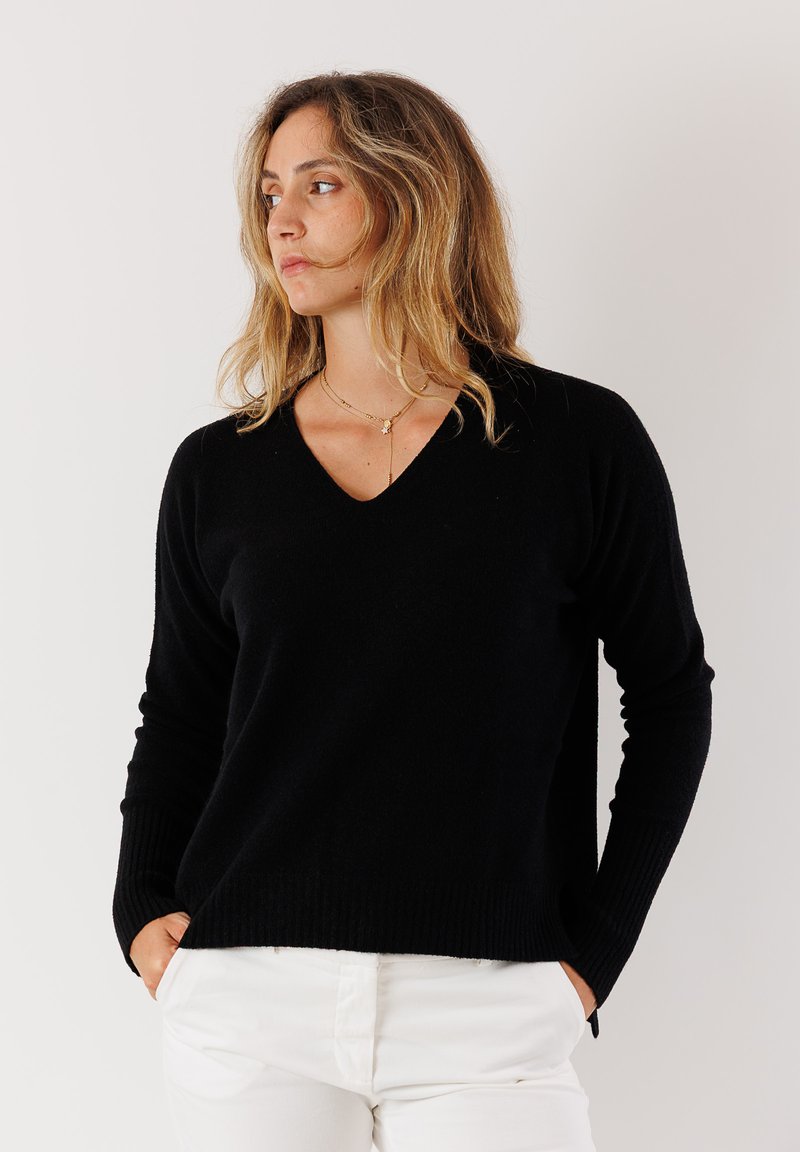 Maglione nero a V con polsini e orlo a costine, realizzato in tessuto morbido. Indossato con pantaloni bianchi, caratterizzati da una vestibilità rilassata e dettagli di cucitura sottili.