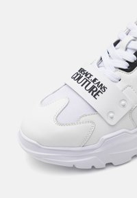 Tenis brancos com sola robusta, apresentando um upper em tecido, detalhes em preto contrastante e uma faixa logo em destaque. Textura suave com detalhes de costura.