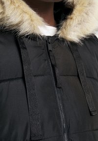 Czarna puchowa kurtka z ozdobnym wykończeniem z sztucznego futra na kapturze, zamek błyskawiczny oraz regulowane sznurki dla pewnego dopasowania. Teksturowany materiał z poziomymi szwami.