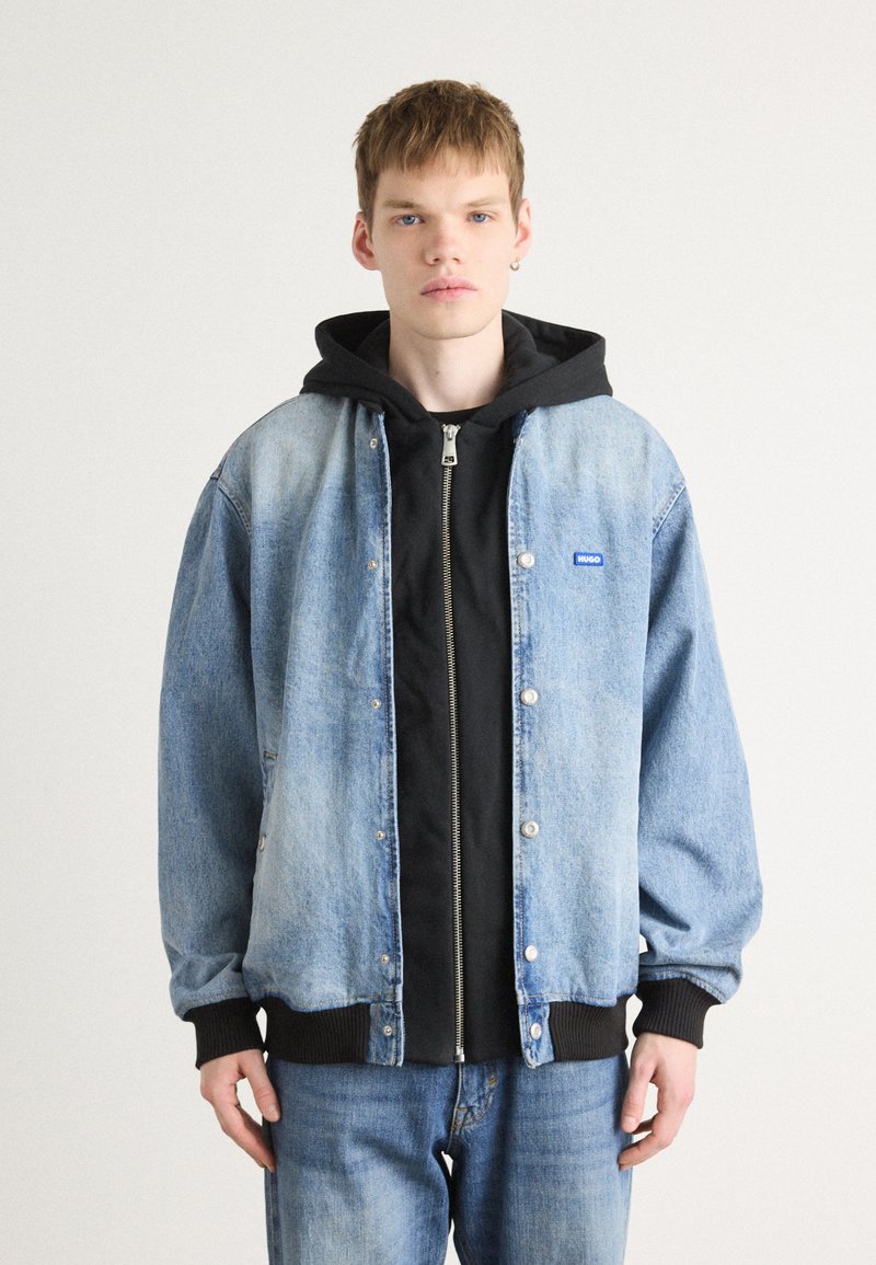 Chaqueta de denim con botones a presión, de un azul claro desgastado, con puños de canalé negros, llevada sobre una sudadera con cremallera negra. El modelo la combinó con jeans oscuros.