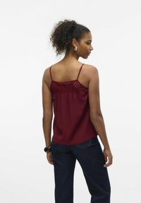 Bordeaux tanktop met kantdetails aan de achterkant, dunne verstelbare bandjes en een losse pasvorm. Gecombineerd met donkerblauwe jeans.