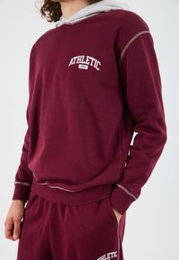 Bordový sveter s šedou kapucňou, s potlačou "ATHLETIC 1997". Mäkká textúra, kontrastné švy a elastické manžety.