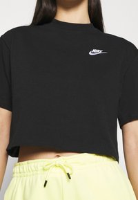 Black cropped T-shirt with short sleeves and a rounded neckline, featuring a white Nike logo on the front. Light yellow sweatpants are visible below.