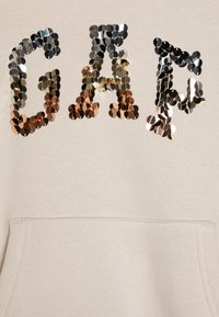 Světlé béžové mikina s flitrovými písmeny "GAP" ve stříbrné, zlaté a měděné barvě. Obsahuje přední klokansko kapsu a žebrované manžety.