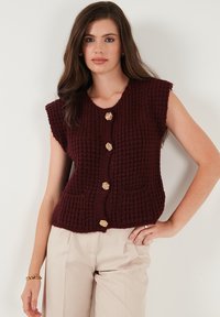 Žena v bezrukávovom tmavo burgundy pletenom vestón s zlatými gombíkmi a béžovými nohavicami, stojaca pred jednoduchým bielym pozadím.