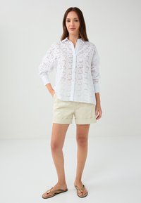 White, button-up shirt with eyelet detailing, long sleeves. Paired with light beige shorts and flip-flops. Smooth fabric texture.