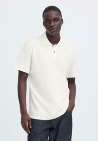 Polo blanc à manches courtes avec col, doté d'une patte à trois boutons et d'une texture subtile. Porté avec un jean en denim foncé.