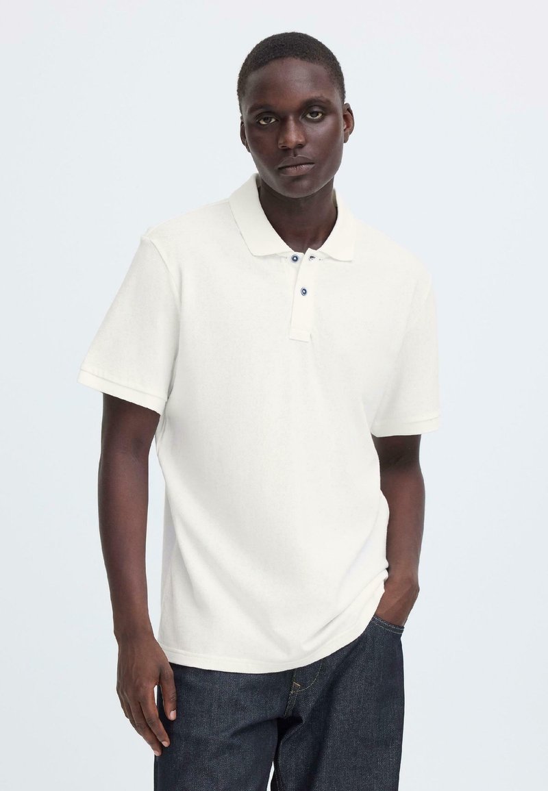 Polo blanc à manches courtes avec col, doté d'une patte à trois boutons et d'une texture subtile. Porté avec un jean en denim foncé.