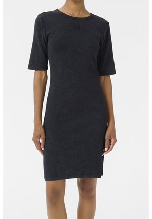 Femme portant une robe noire simple jusqu'aux genoux avec des manches courtes et un col rond, debout sur un fond blanc uni.