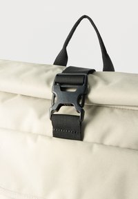 Μπεζ υφασμάτινη τσάντα με μαύρη πλαστική αγκράφα και μαύρο ιμάντα χειρολαβής από νάιλον στο πάνω μέρος, εμφανιζόμενη σε κοντινό πλάνο σε λευκό φόντο.