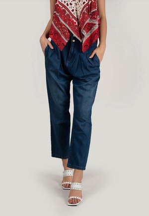 Kvinde iført højtaljede mørkeblå jeans med hænderne i lommerne, en rød- og hvidmønstret top og hvide, nittebesatte, åbne hæle.