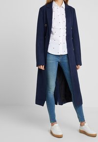Abrigo largo azul marino con dos botones, usado sobre una camisa blanca con pequeños patrones rojos, combinado con jeans azules y zapatillas blancas.