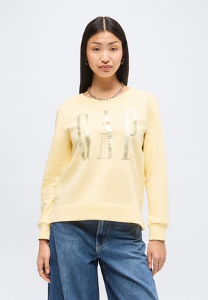 Ung kvinde med langt mørkt hår iført en lysegul GAP-sweatshirt og blå jeans, stående mod en ensfarvet baggrund.