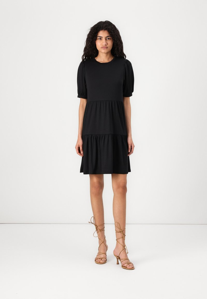 O vestido preto em camadas apresenta um decote redondo e mangas curtas com volume. Feito de um tecido suave. Combinado com sandálias de salto alto em tom bege.