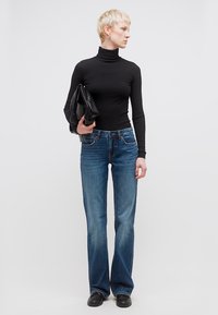 Νέος με κοντά ξανθά μαλλιά, φοράει μαύρο ζιβάγκο, μπλε τζιν, μαύρα παπούτσια και κρατά μαύρο τσαντάκι, κοιτάζοντας προς τα πλάγια.