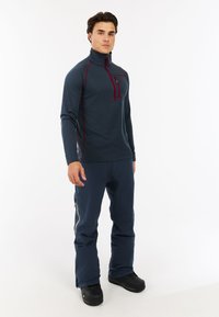 Dunkelblauer Quarter-Zip-Pullover mit einer Brusttasche und kastanienbraunen Akzenten, kombiniert mit navyblauen Hosen. Das Material wirkt glatt und leicht.