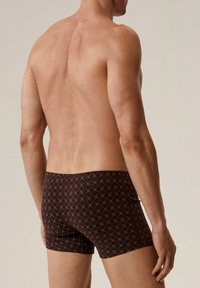 Brązowe kąpielówki w wzory są ozdobione diamentowym wzorem, mają dopasowany krój i niską talię. Materiał wydaje się gładki z minimalną teksturą.