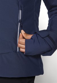 Main tirant sur la fermeture éclair de la poche d'une veste bleu marine, montrant la manche de la veste avec une sangle ajustable en Velcro.