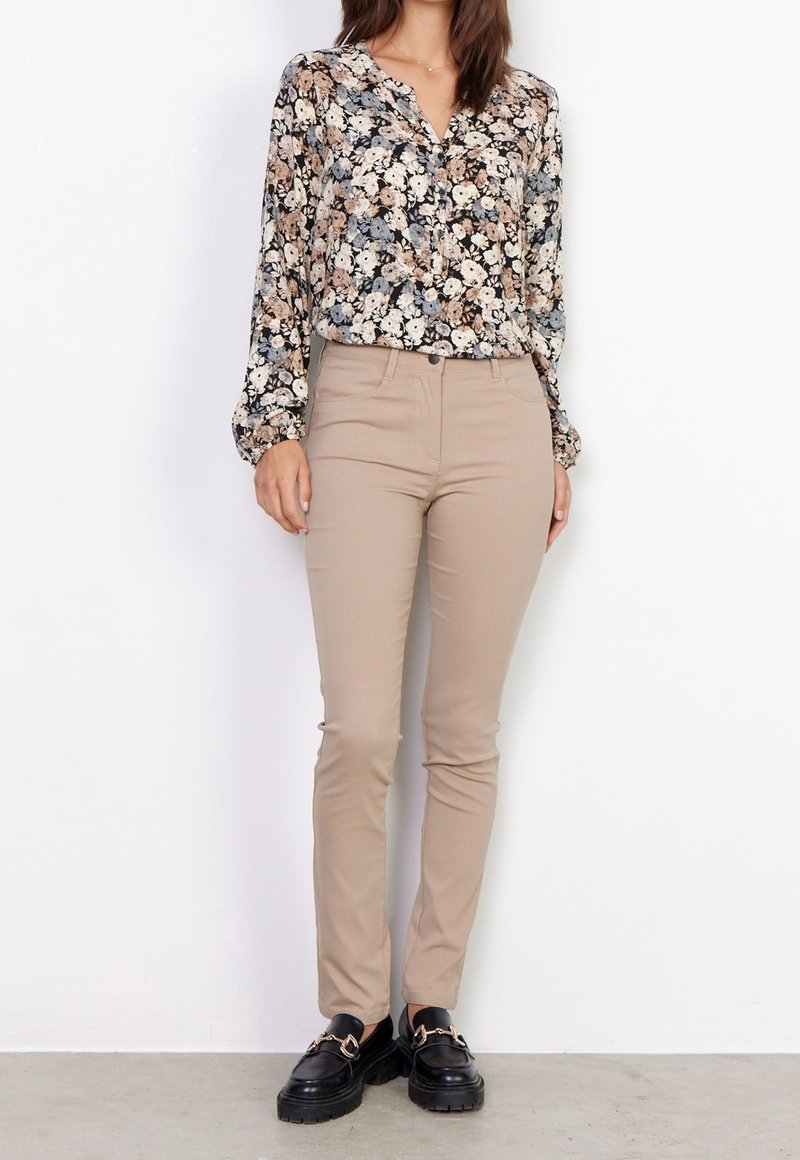 Femme portant un pantalon beige ajusté, un chemisier à manches longues à motif floral, et des mocassins noirs avec détail chaîne dorée, debout contre un mur blanc.