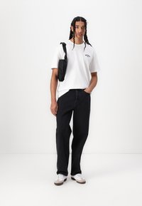 White short-sleeve T-shirt with black logo, paired with loose-fit black jeans. Model wears a small black crossbody bag and white sneakers.