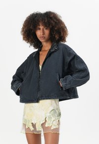 Black cropped jacket with a zip closure and exaggerated sleeves, featuring a textured collar and side pockets, layered over a patterned dress.