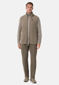 Mees seisab, seljas beež fliisvest heleda pruunika kõrge kaelusega sviidri, pruunid püksid ja beežid spordijalatsid, valge tausta ees.