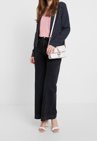 Cardigan bleu marine à pois porté sur un top rose, associé à un jean noir taille haute et des sandales blanches à talons. Accessoires : un sac bandoulière blanc.