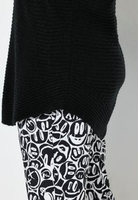 Beine in schwarz-weißen Hosen mit verschiedenen abstrakten Smiley-Gesichtern, kombiniert mit einem strukturierten schwarzen Strickoberteil.