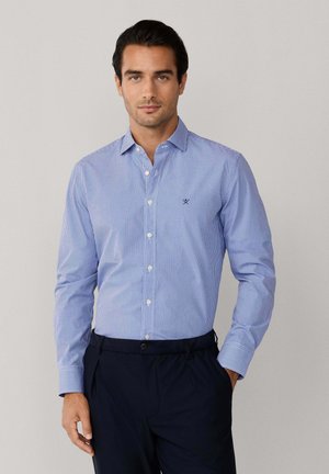 Man die een blauw-wit geruit overhemd met lange mouwen draagt, ingestopt in donkerblauwe broek, staand tegen een effen achtergrond.