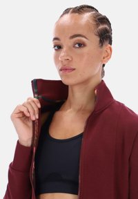 Vínová bunda na zip s vysokým límcem a vzorovaným vnitřkem. Pod ní černá sportovní podprsenka, viditelná texturovaná pokožka.