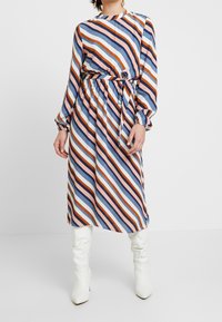 Gestreiftes Midikleid mit diagonalen Streifen in Blau, Orange und Pink, tailliert mit einem Bindegürtel und langen Puffärmeln. Tragen mit weißen Stiefeln.