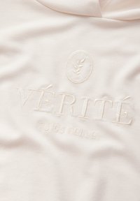 Cremefarbener Stoff mit einem geprägten Design, das den Text "VÉRITÉ" und "TU ES BELLE" in einem dezenten, strukturierten Stickstil zeigt.