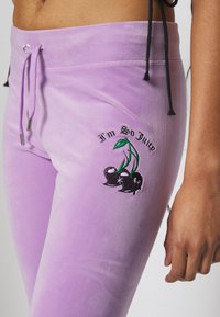 Violeta sporta bikses no mīksta auduma ar aukliņu jostasvietā, sānu kabatām un izšutu ķiršu dizainu ar tekstu "Es esmu tik sulīgs."