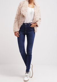 Plufsig beige jacka med sjalkrage, bärs över en vit blus, ihop med mörkblå skinny jeans och vita snörskor.