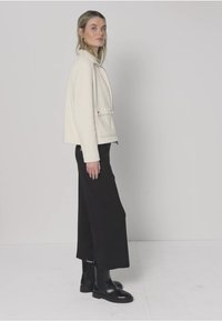 Chaqueta color crema con un diseño estructurado, que presenta un cuello y bolsillos delanteros, combinada con pantalones negros de pierna ancha y botas negras.