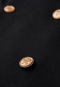 Tissu noir au toucher lisse avec un bouton doré, orné d'un blason et de bords décoratifs, placé de façon décentrée.